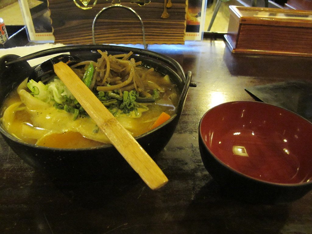 木桌上一個黑色鐵鍋裡的餺飥(ほうとう):味噌湯裡有寬扁的麵、南瓜、紅蘿蔔、青菜和山菜,上面架著一根木杓;右邊放著一個內側紅色的空碗