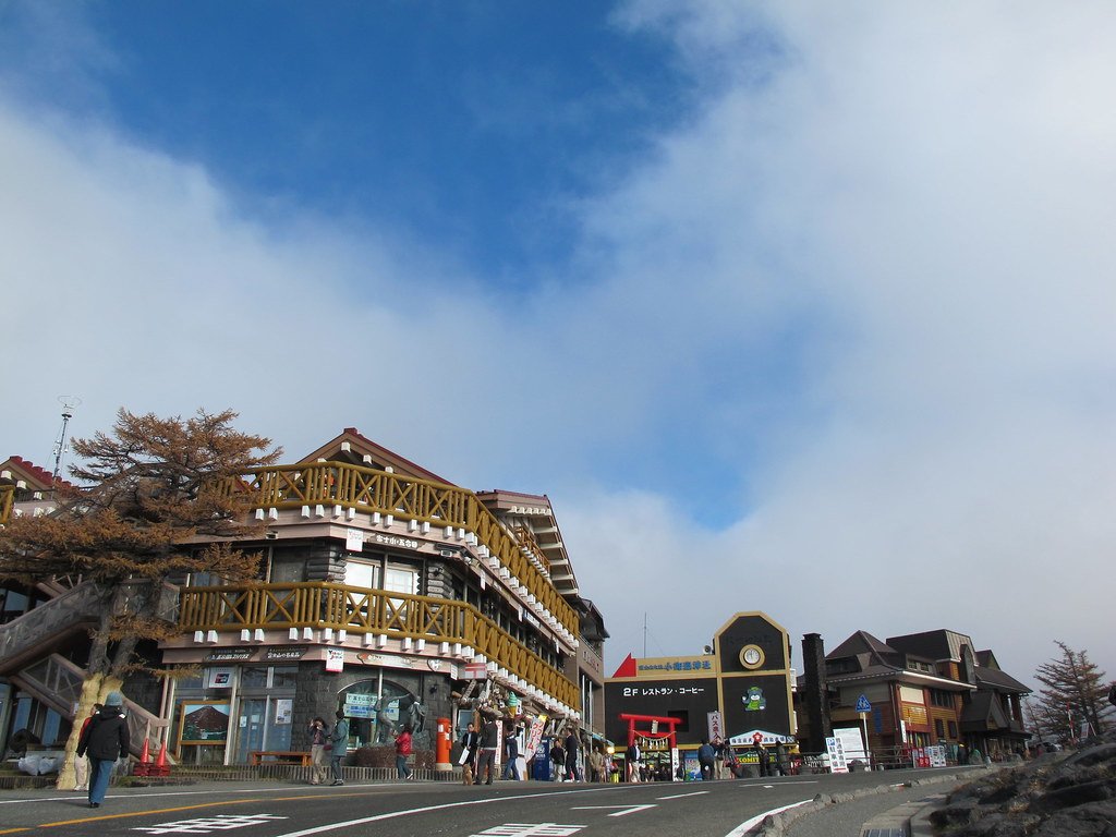 富士スバルライン五合目的店家一條街:左邊是有木頭陽台的兩層樓賣店,旁邊一棵轉黃的落葉松,中間是掛著時鐘、寫著「2F レストラン・コーヒー」的建築與紅色鳥居,右邊還有幾棟山屋式的房子;天空一半藍天一半雲,路上有零星遊客