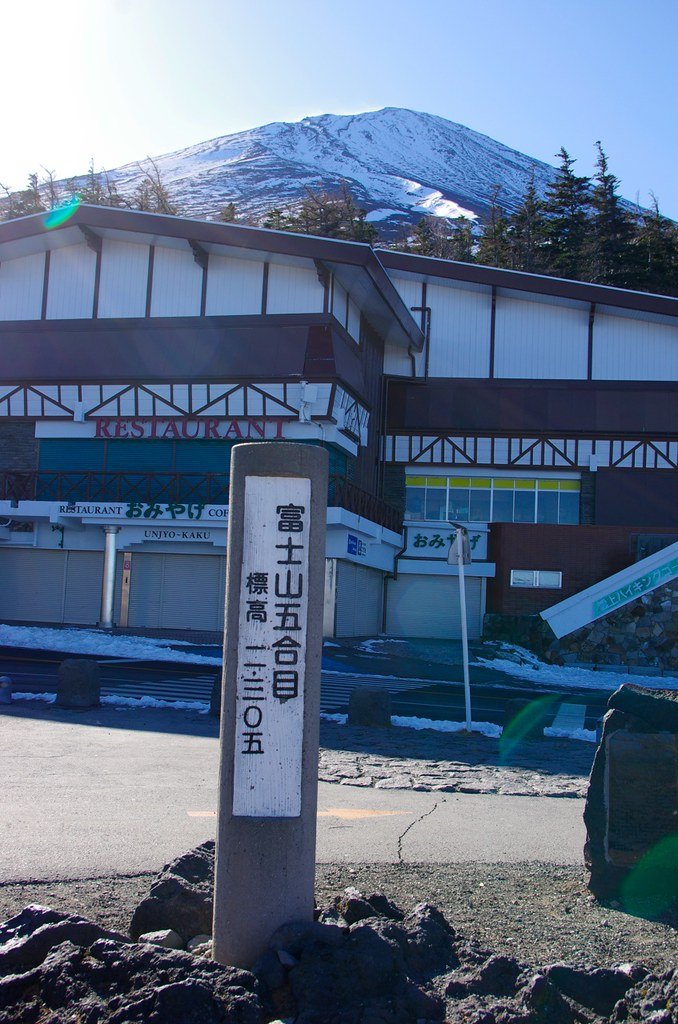富士スバルライン五合目寫著「富士山五合目 標高二三〇五」的石柱,後方是雲上閣的建築,招牌寫 RESTAURANT、おみやげ、UNJYO-KAKU,一樓的鐵捲門全部拉下;地上有殘雪,再後方是山頂積雪的富士山與藍天