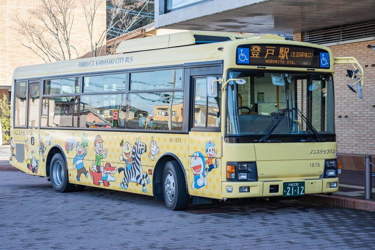 停在博物館門口的黃色川崎市巴士，車身畫滿藤子・F・不二雄的角色，車頭的橘色跑馬燈寫著「登戸駅（生田緑地口）NOBORITO STA.」，車側寫著「川崎市バス KAWASAKI CITY BUS」