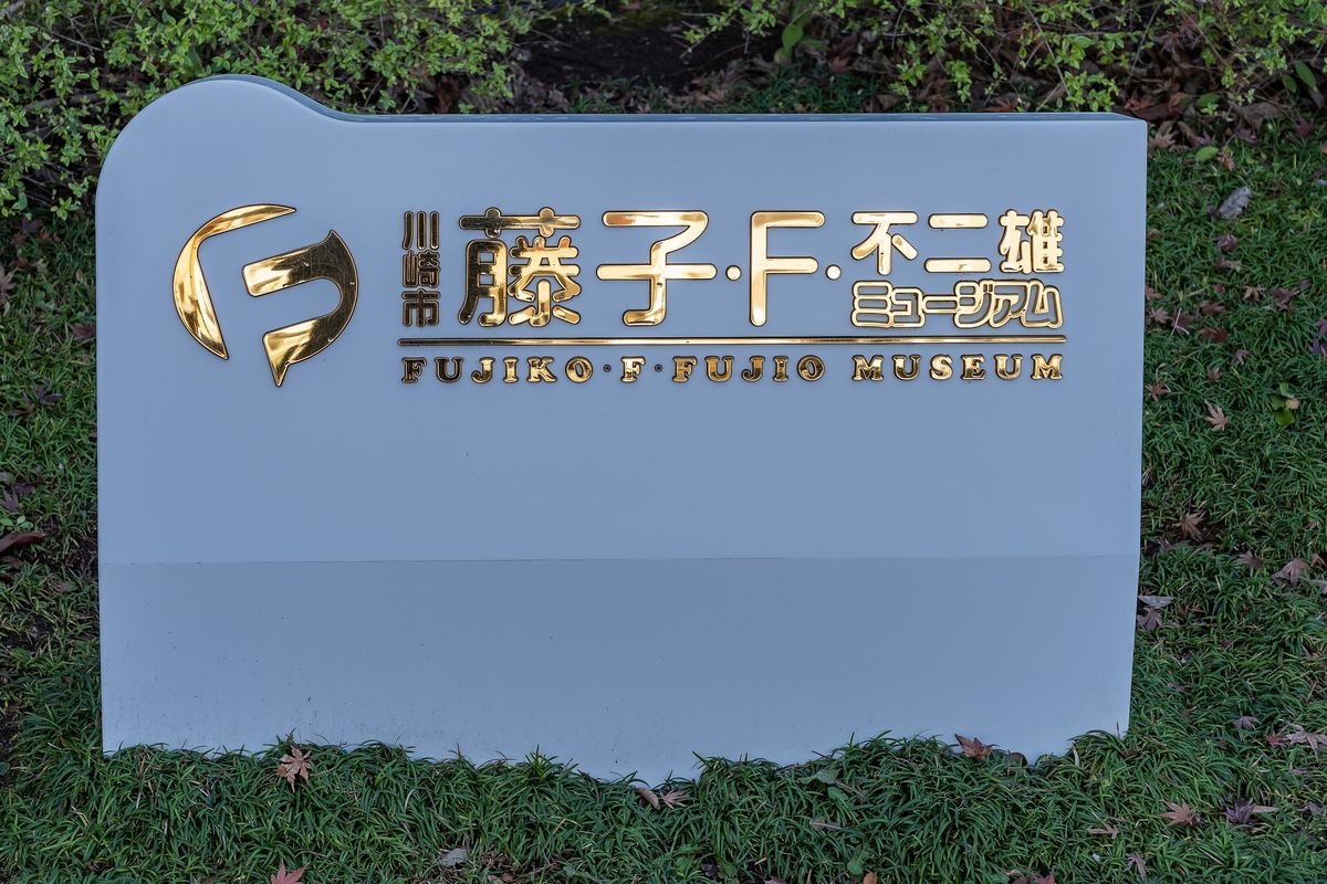 草地上一塊淺灰色石材館名牌，金色立體字寫著「川崎市 藤子・F・不二雄ミュージアム」與英文 FUJIKO・F・FUJIO MUSEUM，左邊是金色的 F 字標誌，背後是灌木叢