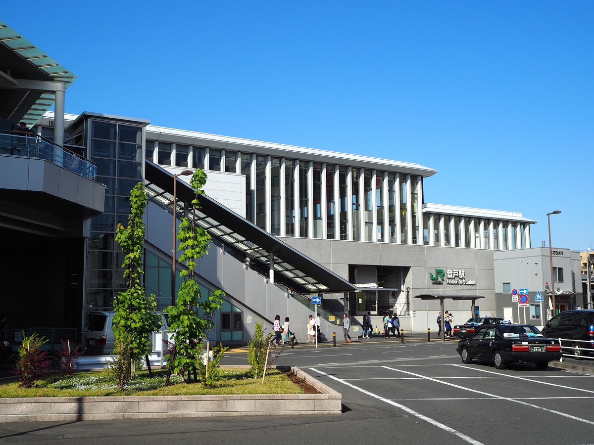 晴天的 JR 登戶站站房，灰色外牆上有綠色 JR 標誌與「登戸駅 Noborito Station」字樣，上方是一整排白色直條窗；左邊一道長長的電扶梯與樓梯從天橋下到地面，站前是計程車停靠的廣場，右邊有一間寫著 KOBAN 的派出所