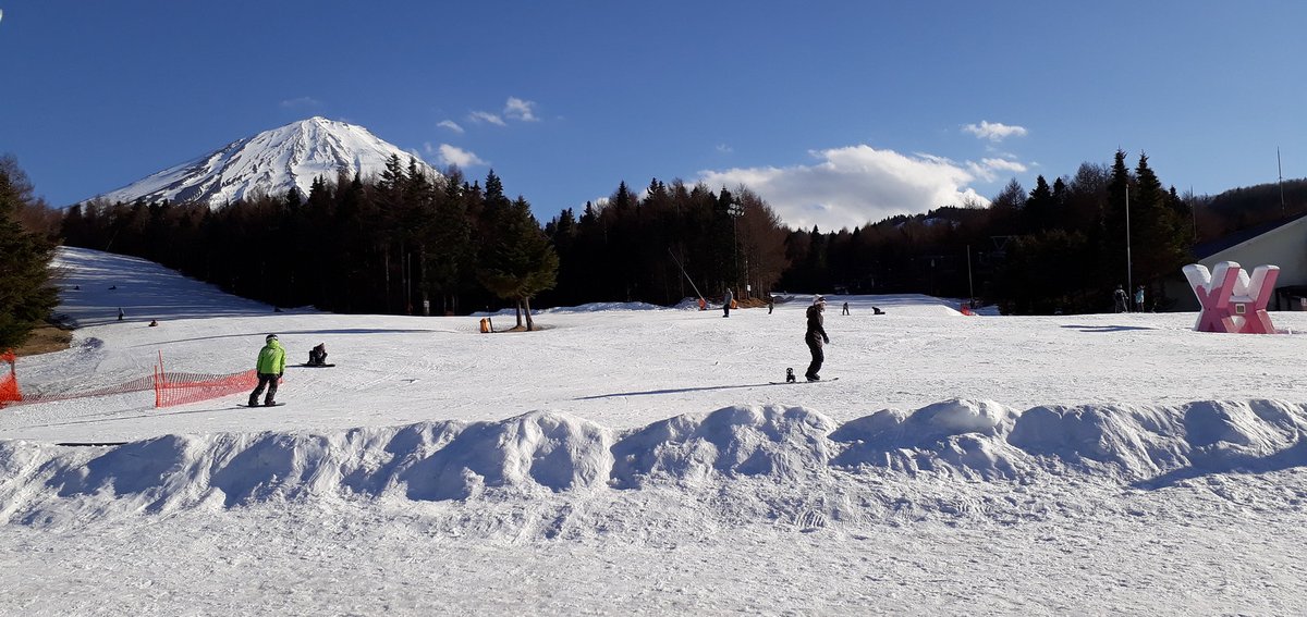 富士天山腳一片寬闊平緩的雪坡,前景是一道推起來的雪堆,雪坡上幾個單板滑雪者,一人穿螢光綠外套,左邊拉著橘色安全網,右邊有粉紅色的大型立體字裝飾;遠處針葉林上方是積雪的富士山,左邊一條雪道沿樹林往上;2017 年 3 月 10 日攝
