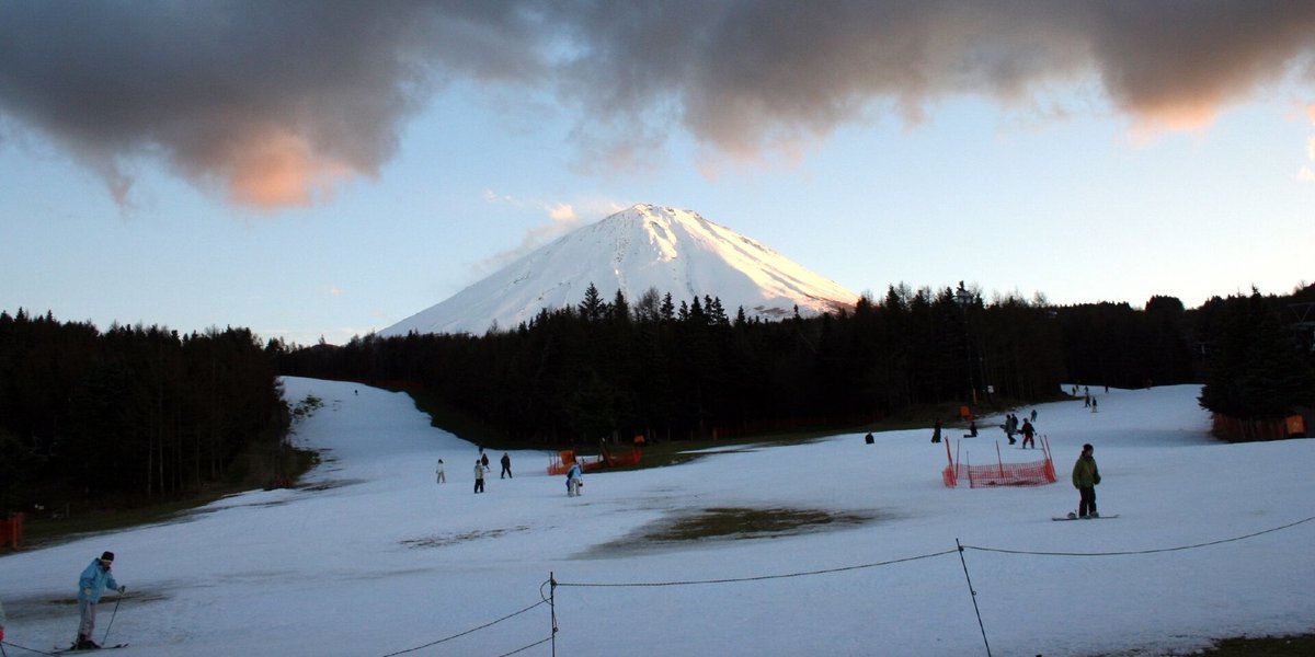 黃昏時的富士天山腳:遠方富士山頂被夕陽照成淡橘色,前景雪面很薄,中間露出一塊褐色草地,左邊一條窄雪道往上通進樹林,幾個人在雪上滑行,近處拉著一條低矮的繩子;2006 年 12 月 27 日攝