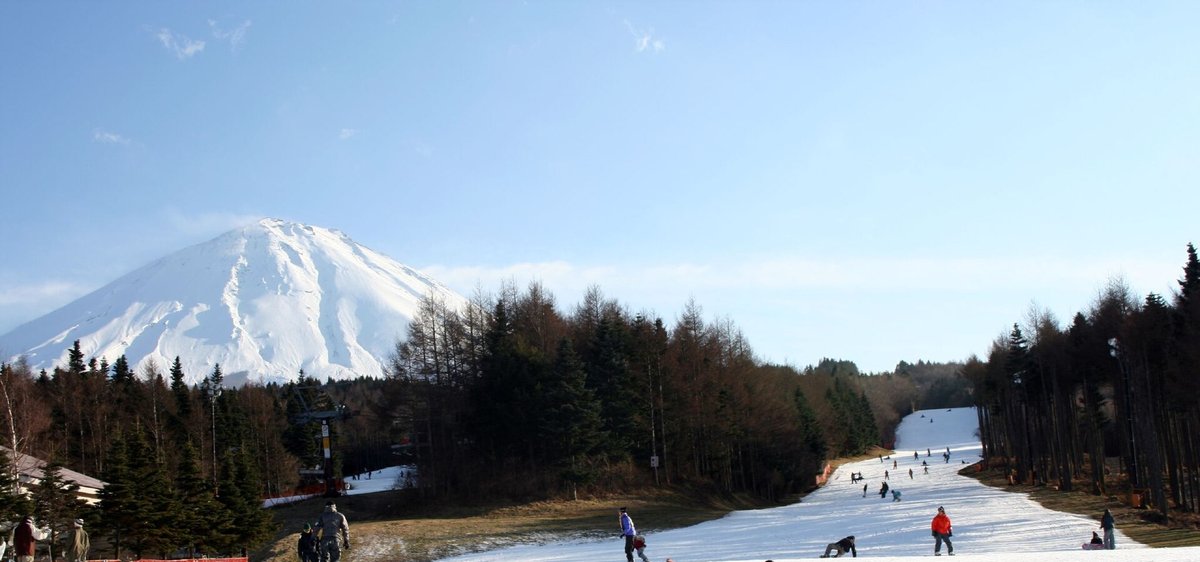 晴天的富士天:左上是積雪的富士山,右邊一條白色雪道從林間延伸下來,雪道外的坡地是褐色枯草、沒有積雪,雪道上散著滑雪和單板的人;2006 年 12 月 27 日攝