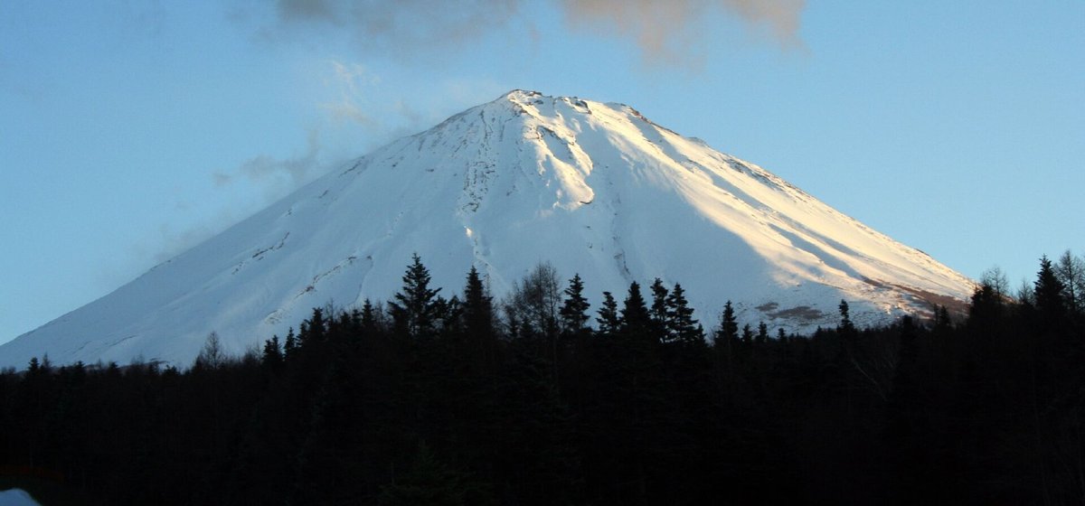 傍晚的富士山,山頂與右半邊山坡被夕陽照成淡金色,上方壓著一大片灰色雲層,山腳是一整排剪影般的黑色針葉林;2006 年 12 月 27 日攝於富士天滑雪場
