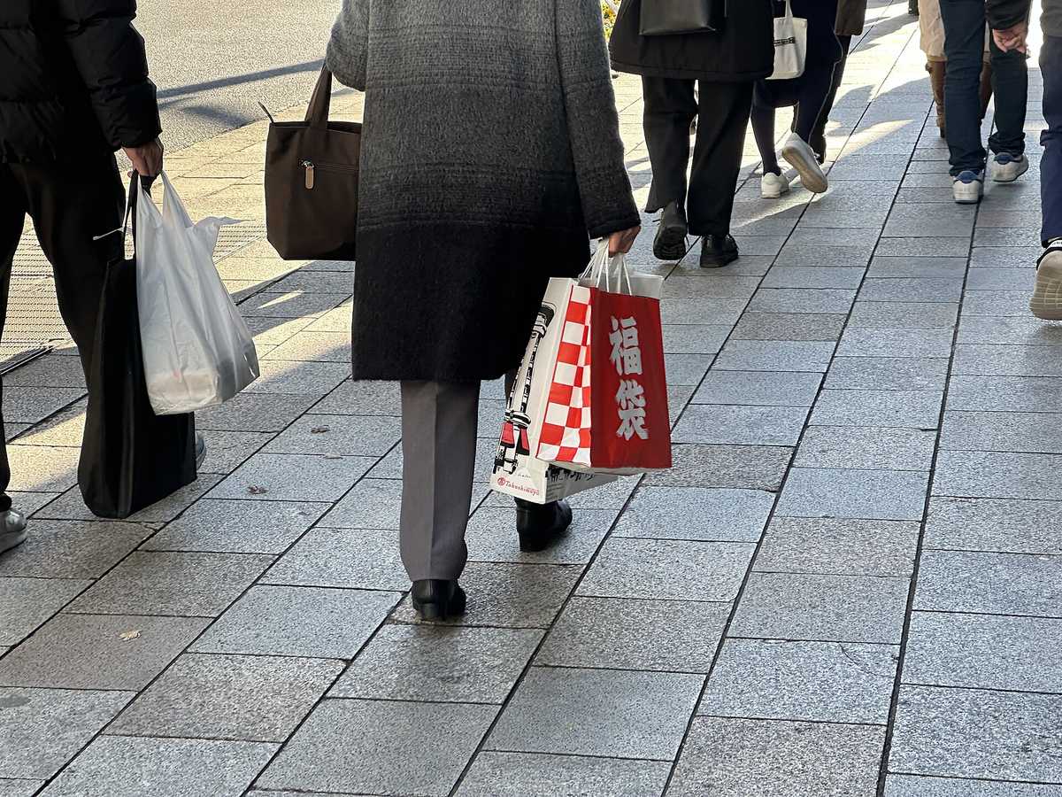 Shoppers on a Tokyo pavement carrying a red fukubukuro lucky bag, framed from the waist down