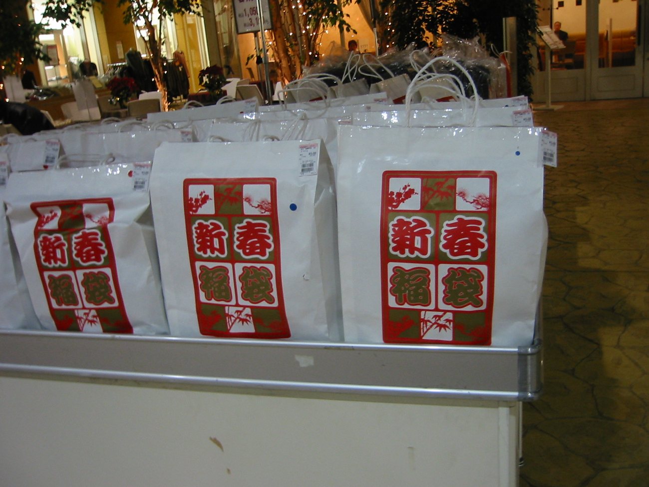 A shopfront table stacked with white paper lucky bags printed with a red and green shinshun fukubukuro design, each carrying a price tag