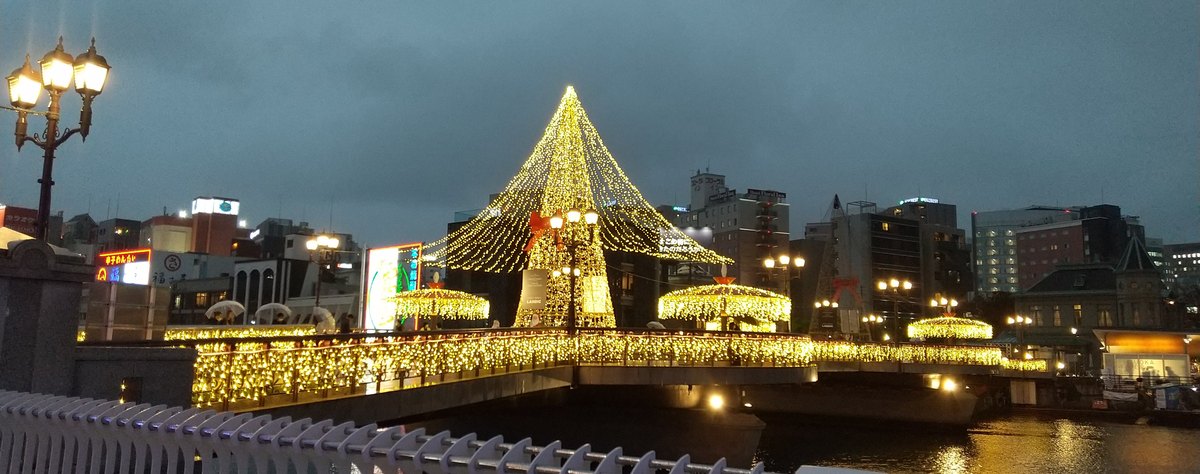 2022 年 11 月陰雨的傍晚隔著那珂川看福博であい橋：整座橋的欄杆掛滿黃色燈串，橋中央一座綁著紅色蝴蝶結的黃色燈錐，兩旁各有一座燈傘，橋上有撐傘的行人，左前方是復古造型的街燈，右側看得到貴賓館的尖塔屋頂