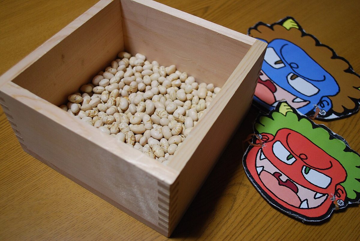 Roasted soybeans (fukumame) in a wooden masu measure beside red and blue paper oni masks, the standard Setsubun kit sold in Japanese supermarkets