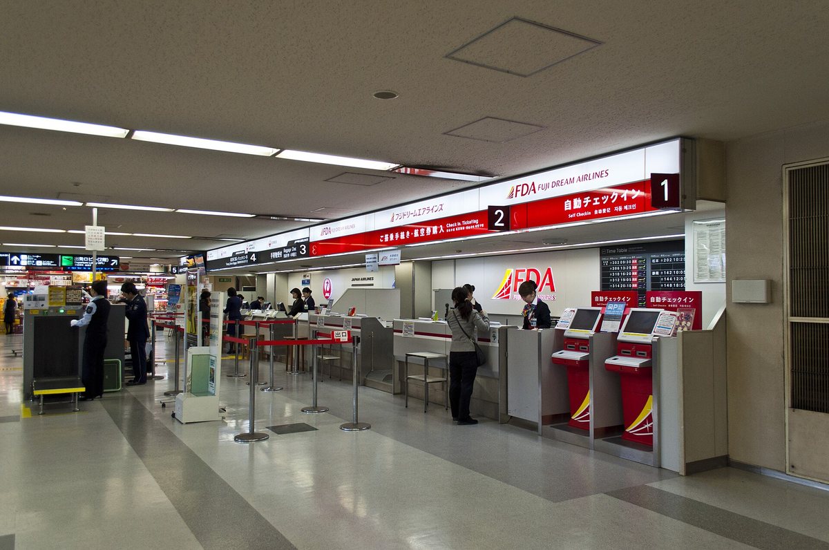 A domestic check-in area at Fukuoka Airport, a red illuminated sign reading 自動チェックイン Self Checkin in Japanese, English, Chinese and Korean above counters numbered 1 and 2, two red self check-in kiosks on the right, a staffed Japan Airlines counter with the crane logo on the left