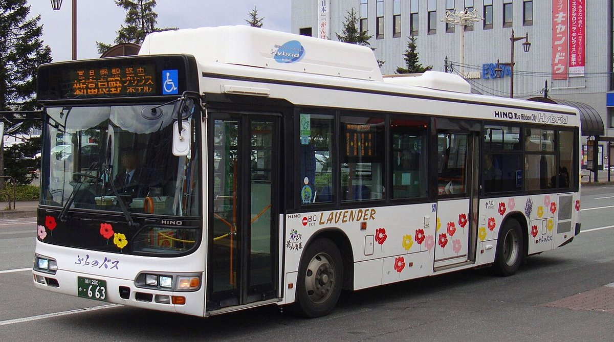 停在路邊的白色ふらの巴士,車身畫著紅、黃、粉紅色的花與「LAVENDER」字樣,車頭橘色電子方向幕顯示「富良野駅経由 新富良野プリンスホテル」,背景是灰色的商業大樓
