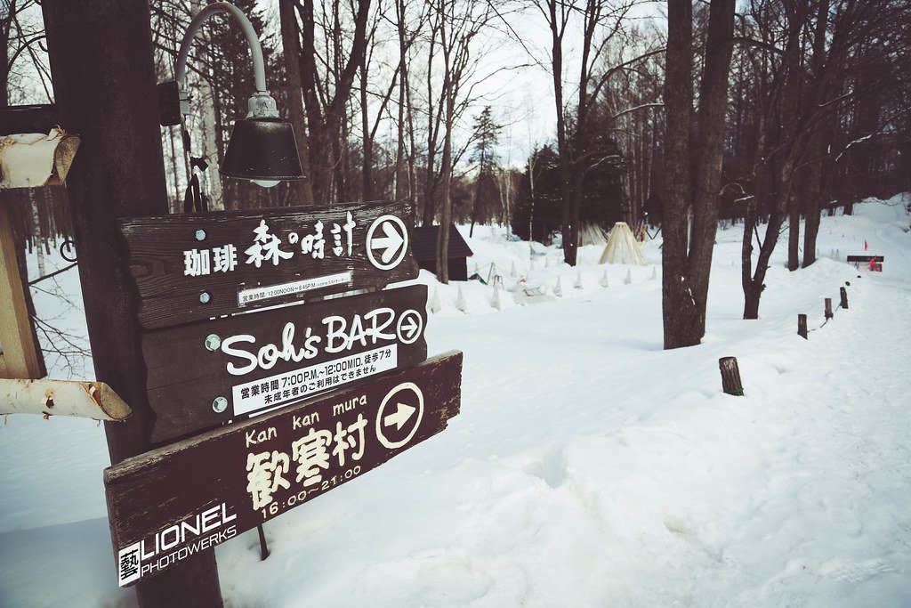 雪地樹林邊的木柱上釘著三塊深色木牌,由上到下寫著「珈琲 森の時計」「Soh's BAR」「Kan kan mura 歓寒村 16:00–21:00」,箭頭都指向右邊;右後方樹林間有一頂圓錐形帳篷和幾排小雪堆