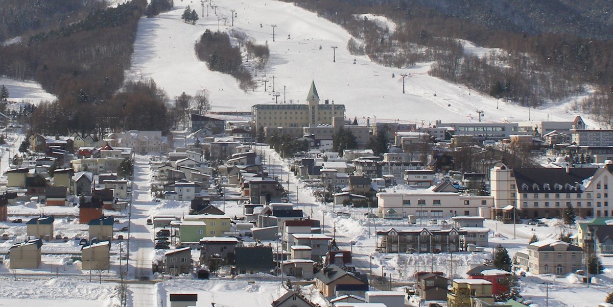 北之峰雪道山腳的旅館街:畫面上半是寬闊的雪道、吊椅支柱與空中吊籃車廂,雪道底緊鄰一棟有綠色尖塔的米黃色旅館,往下是幾條積雪的街道與成排的小型旅館、民宅