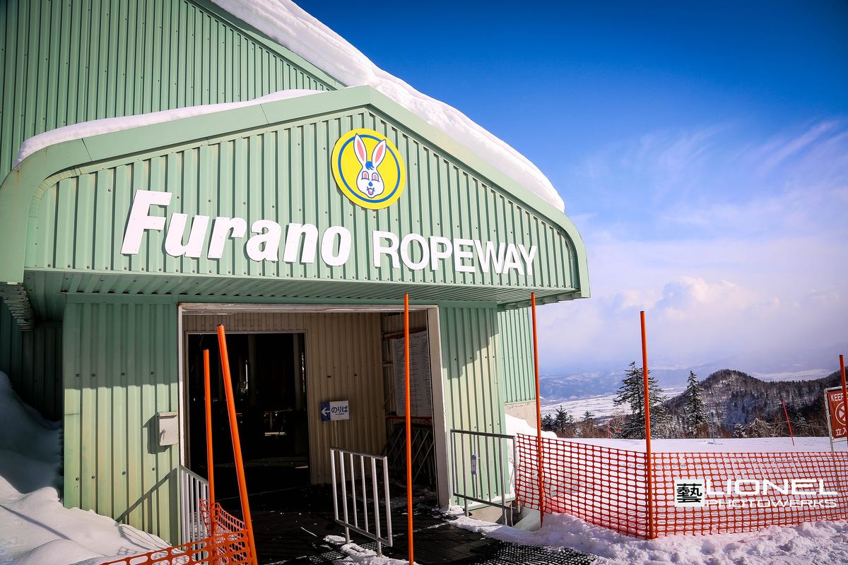 富良野纜車山頂站:屋頂積著厚雪的綠色鐵皮建築,門楣上有兔子標誌與「Furano ROPEWAY」字樣,門口立著「のりば」箭頭牌,出口外圍著橘色的網,右邊遠處看得到山下的盆地