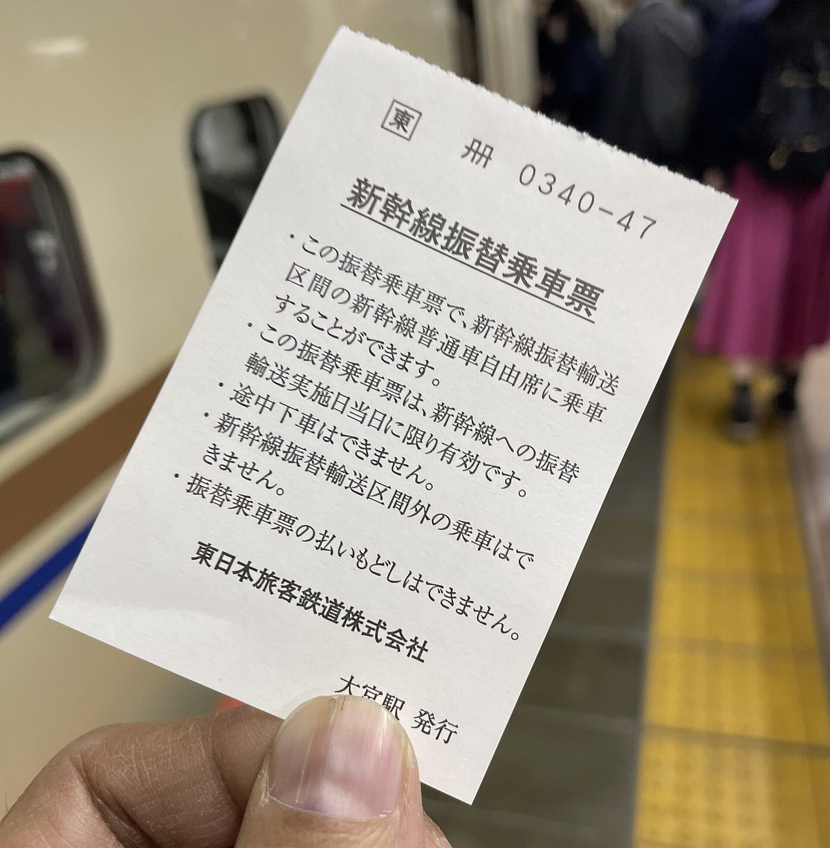 A hand holding a JR East shinkansen substitute travel slip issued at Omiya Station, its printed conditions listing same-day validity, no stopovers, no travel outside the section and no refunds, with a shinkansen at the platform behind