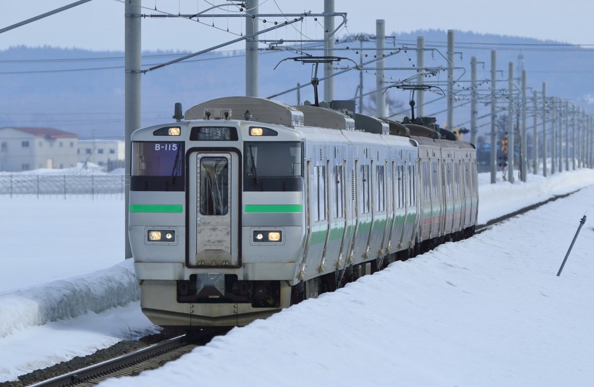 JR 北海道 733 系電車行駛在學園都市線上，車頭編號 B-115，兩側鐵軌與田野都被厚雪覆蓋，背景是電線桿與遠山