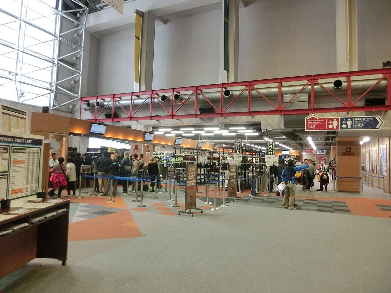 Indoor rental reception hall at GALA Yuzawa ski resort, with a RENTAL PRICE LIST board listing separate SNOWBOARD and SKI columns and a queue of visitors behind blue belt barriers