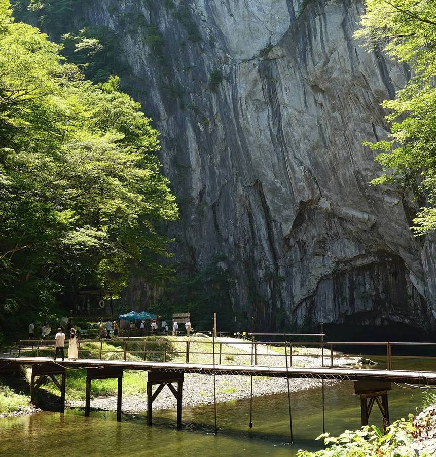 大猊鼻岩前的折返點:一座灰白色的巨大岩壁佔滿畫面,底部有一個凹進去的岩洞,岩壁前是一片碎石河灘,河灘上有藍色帳篷與一群遊客,前景一座有欄杆的鐵架便橋橫過溪水,兩位遊客正走在橋上