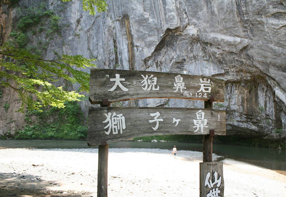 大猊鼻岩下的木製指示牌:上排寫「大猊鼻岩 高さ124m」,下排寫「獅子ヶ鼻」,背後是灰白色的大岩壁,岩壁下有一片白色碎石河灘與溪水