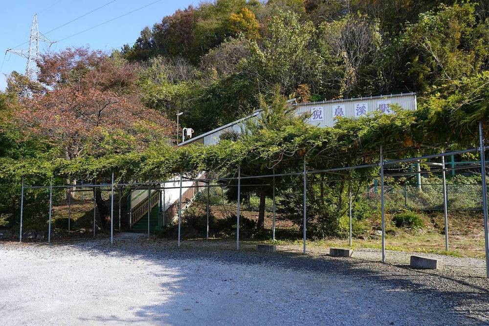 JR 大船渡線猊鼻渓站外觀:一棟架高的白色鐵皮站房,牆上寫著「猊鼻渓駅」,左邊一道綠色樓梯通上月台,站前是碎石空地與爬滿藤蔓的棚架,背後山坡的樹葉開始轉紅
