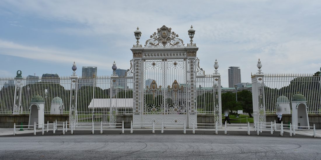 迎賓館正門:一座白色鑄鐵大門,門頂有金色徽章與捲草裝飾、兩側各一盞燈,左右連著一整排白色鐵柵欄,柵欄前各有一座綠色圓頂的白色崗亭,最前面是一排白色矮柱;透過柵欄看得到裡面的松樹和遠處本館的綠色屋頂,天空多雲,柵欄內有兩位工作人員走過