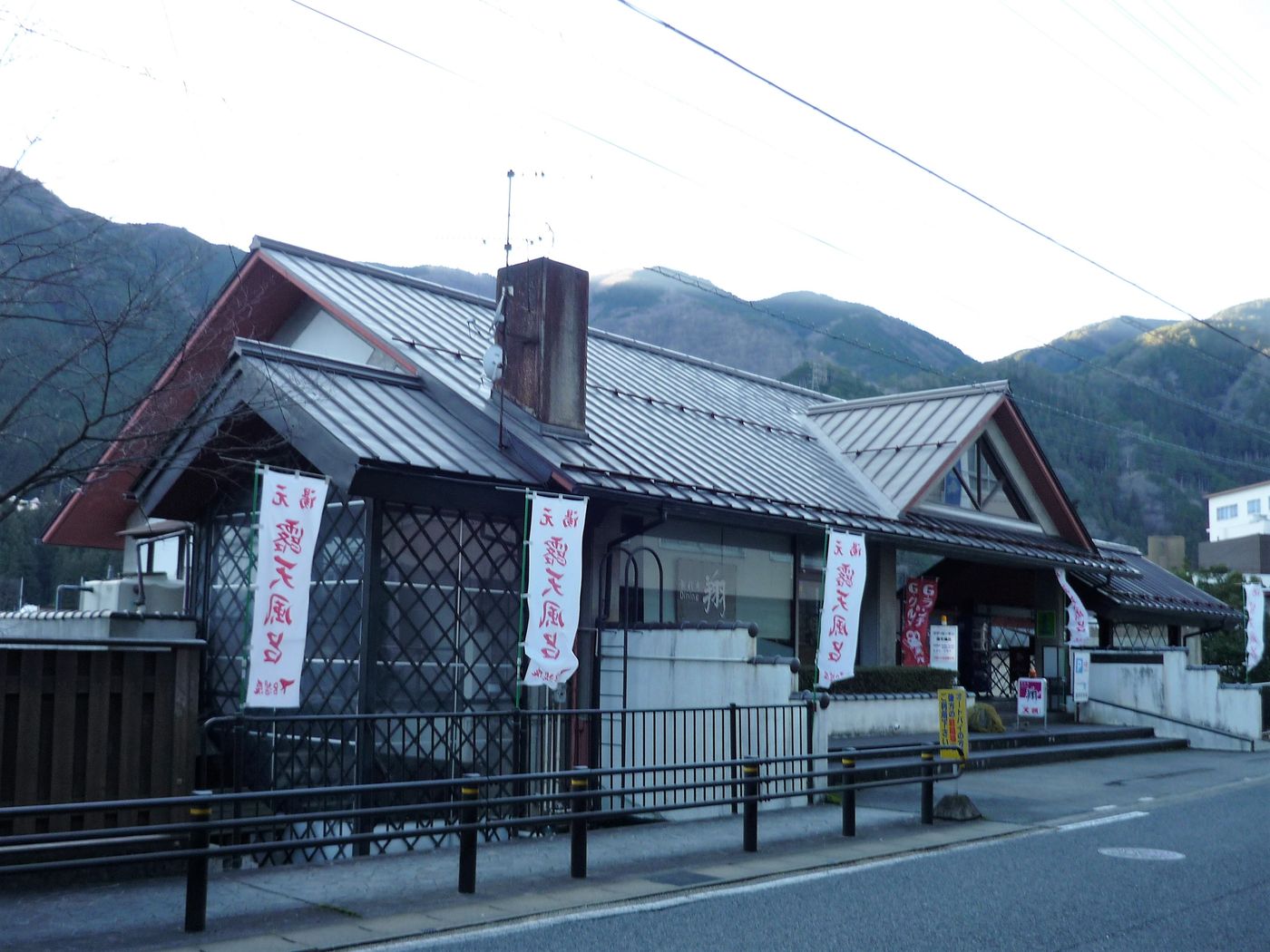 クアガーデン露天風呂的入口:瓦頂的和風平房,門前立著幾面寫著「露天風呂」的白色旗幟,背後是山