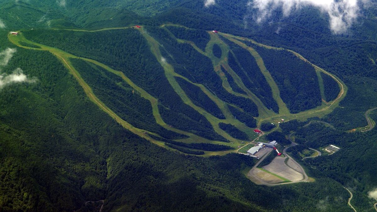 夏天從空中俯瞰夏油高原滑雪場:深綠色森林裡切出十幾條淺綠色的草地條帶,從山頂往下呈扇形散開、在右下方匯集,匯集處有幾棟白色與紅色屋頂的建築和一大片停車場