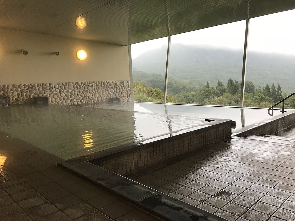 兎森の湯的大浴池:沒有人,貼著磁磚的方形浴池,左邊牆面下半是鵝卵石,牆上兩盞圓形壁燈,右邊整面落地開口望出去是長滿綠樹的山坡和雲霧