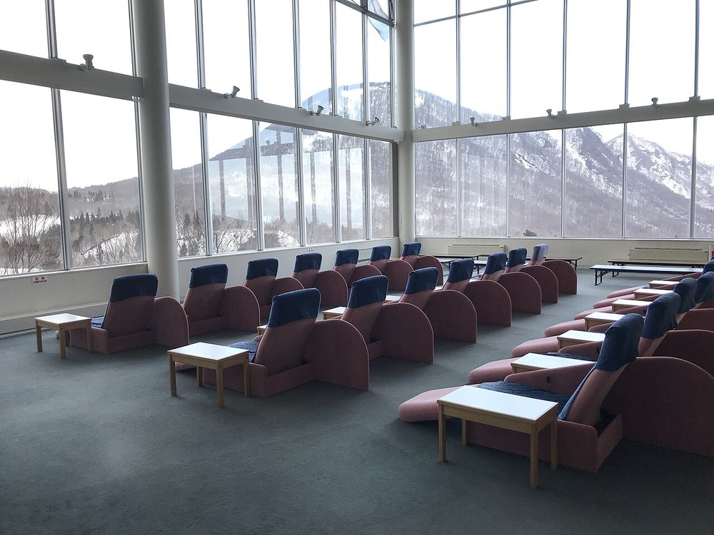 挑高的休息室:兩面落地玻璃窗外是覆雪的山,地上鋪綠灰色地毯,排著二十幾張粉紅色底、藍色椅背的躺椅,每兩張之間有一張白色小桌,沒有人