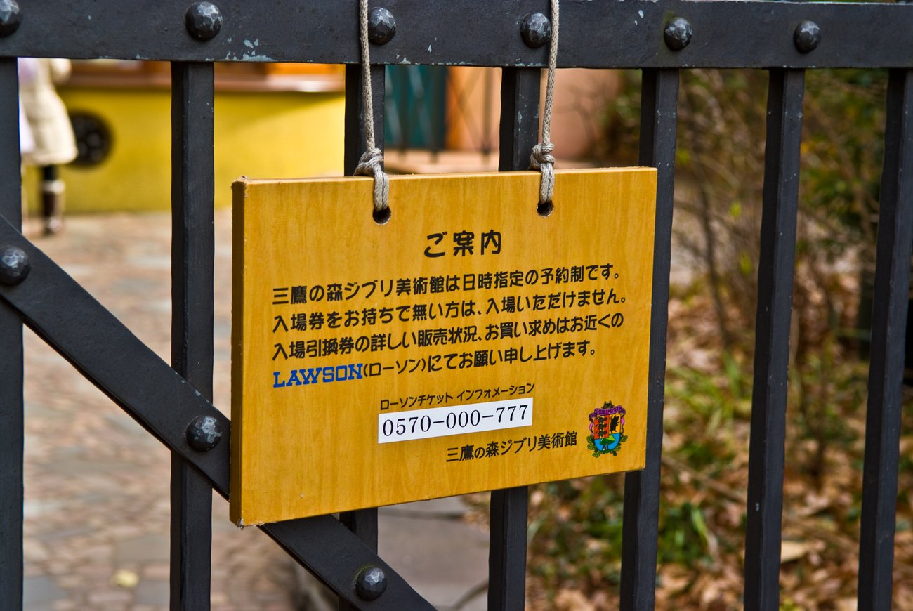 A tan wooden notice board hung by two lengths of twine from a black wrought-iron gate; the board is headed ご案内 in black, with four lines of Japanese stating that the museum runs on date-and-time-designated reservations, that visitors without an admission ticket cannot enter, and that sales and availability should be checked at a nearby LAWSON, the word LAWSON printed in blue; below it a boxed telephone number 0570-000-777, and at lower right the museum's colour crest beside the signature 三鷹の森ジブリ美術館; behind the gate a blurred yellow wall and flagstone paving