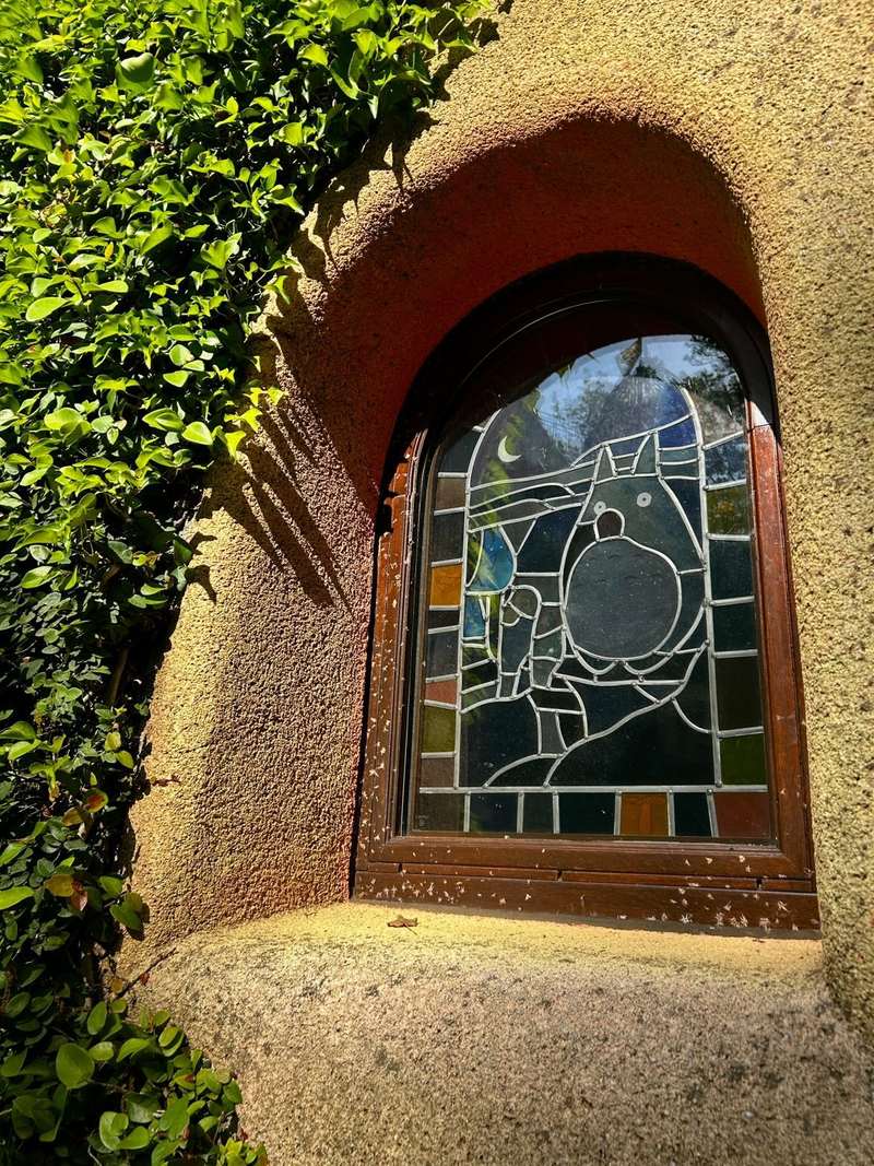 An arched wooden-framed window set into a sunlit cream-coloured rough stucco wall; the leaded stained glass shows a round grey-blue Totoro with pointed ears in deep green and blue glass, a smaller figure beside it and a white crescent moon, over a border of amber and green rectangular panes; the glass reflects the trees and sky above, white blossom fluff and a dead leaf lie on the frame and sill, and ivy climbs the wall on the left