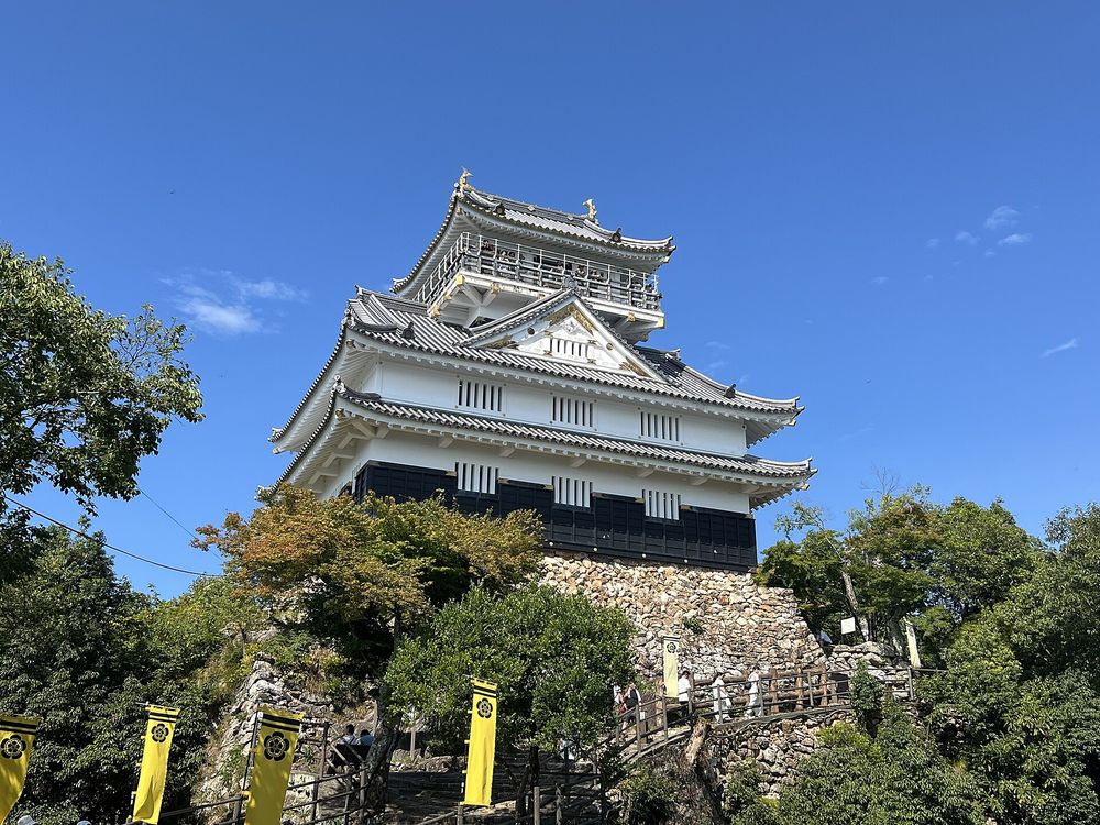 整修前的岐阜城天守（2024 年 8 月）：藍天下白牆黑色腰板的三層天守立在石垣上，頂層有一圈觀景迴廊，前方步道兩側插著黃底黑色家紋的旗幟