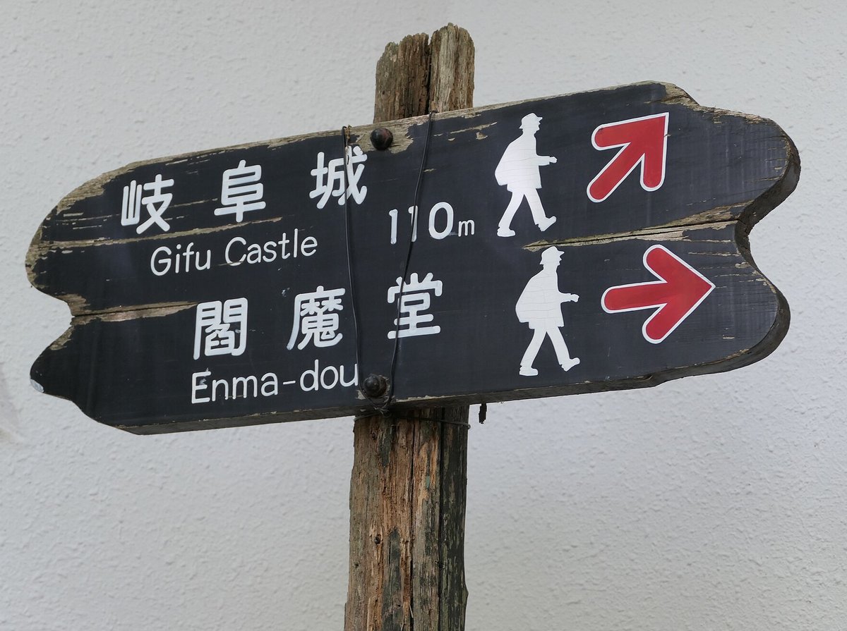 金華山山頂的黑色木製路標：上排寫「岐阜城 Gifu Castle 110m」配右上紅色箭頭，下排寫「閻魔堂 Enma-dou」配向右紅色箭頭，兩排都畫著白色步行人形