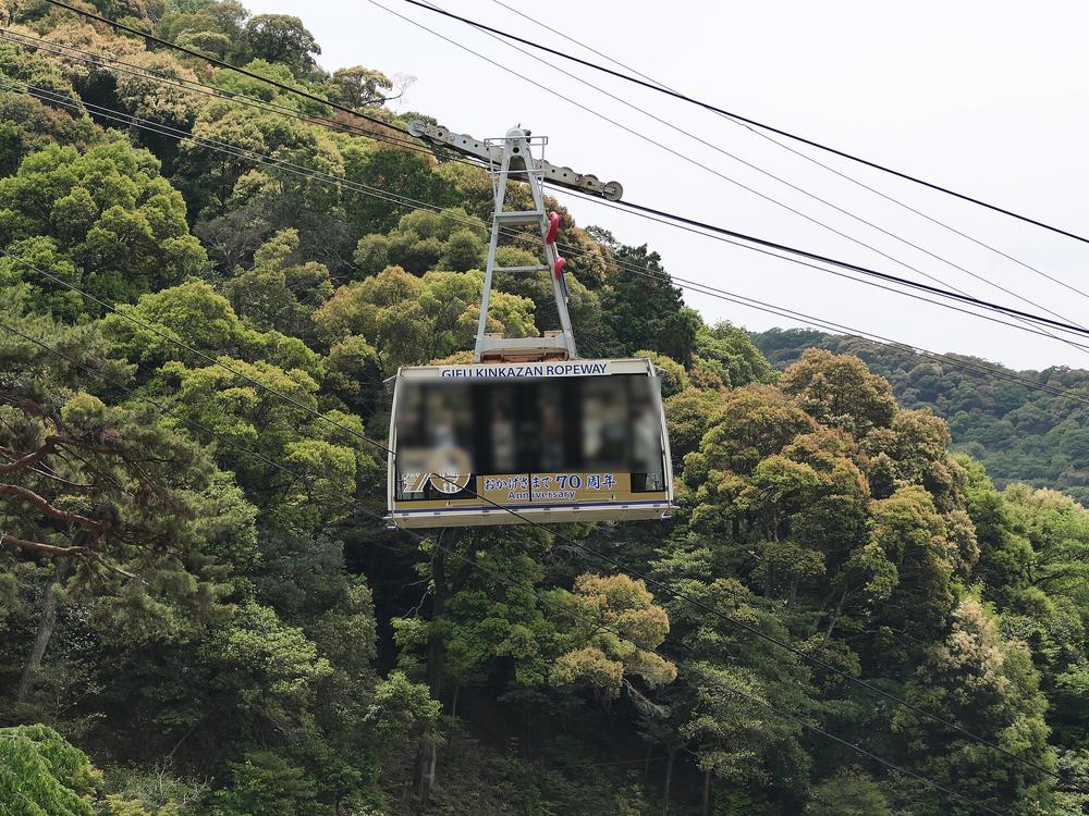 金華山纜車車廂懸在纜索上，背景是整片綠色山林；車廂上方寫著 GIFU KINKAZAN ROPEWAY，下方是「おかげさまで 70 周年」的標示