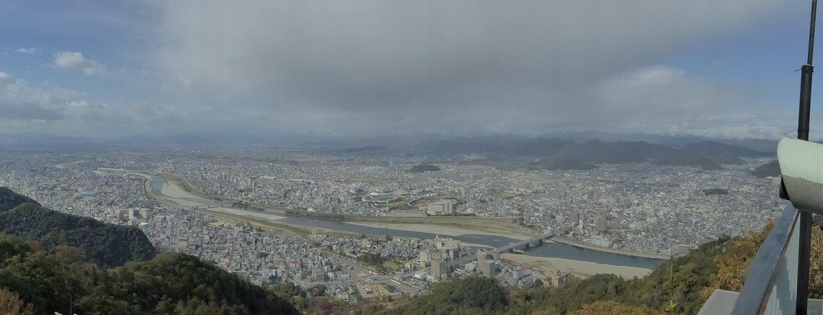 從金華山展望台往北看的全景：長良川從左往右彎過岐阜市區，河上有兩座橋，河岸是大片白色礫石灘，遠方是一排山脈，右緣看得到展望台的欄杆和投幣望遠鏡