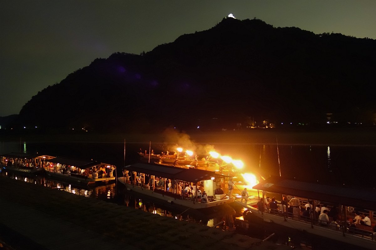 夜晚的長良川鵜飼：河面上幾艘鵜舟的篝火燒得通紅、冒著煙，旁邊停靠著坐滿遊客的屋形觀覽船，背後是漆黑的金華山，山頂有一點打了燈的天守