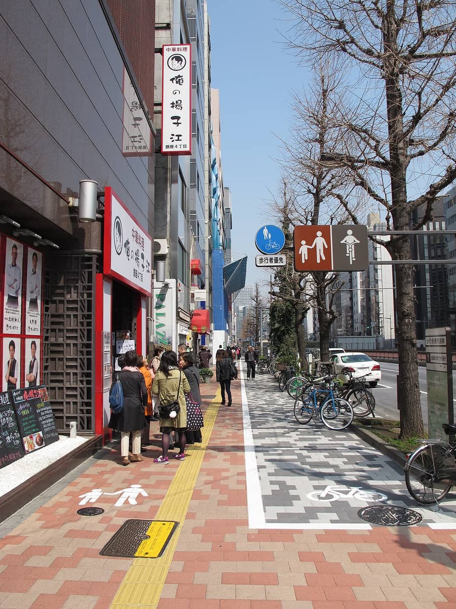 A footway in Ginza 4-chome, Tokyo: a round blue sign showing an adult holding a child's hand above a bicycle, with a white plate beneath it reading pedestrian priority, next to a brown two-panel pictogram with pedestrians on the left and a cyclist on the right; the paving is split by a white line, red brick with a pedestrian symbol on the left where people queue outside a restaurant, grey patterned paving with a bicycle symbol on the right, next to the carriageway