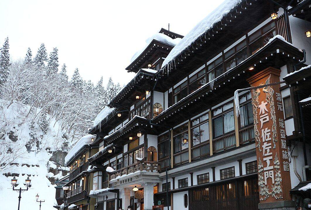 雪中的能登屋旅館:三層木造建築的屋簷壓著厚雪、垂著冰柱,走廊一排吊燈亮著,右側立著刻有鳳凰浮雕的「木戸佐左エ門」看板與「湯元 能登屋旅館」直式招牌