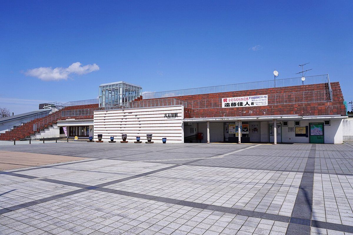 JR 大石田站西口站舍:紅磚色斜屋頂搭白色橫條牆面,牆上寫著「大石田駅」,站前是一大片空曠的方磚廣場