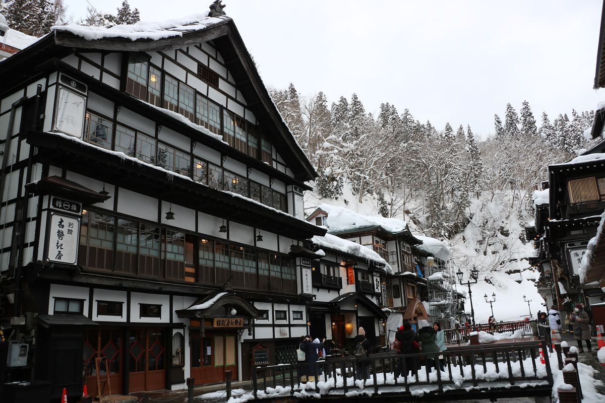 Ginzan Onsen in winter: the three-storey wooden Kosenkaku Bekkan on the left with snow and icicles on its eaves, facing more ryokan and a snow-laden cedar slope across the river