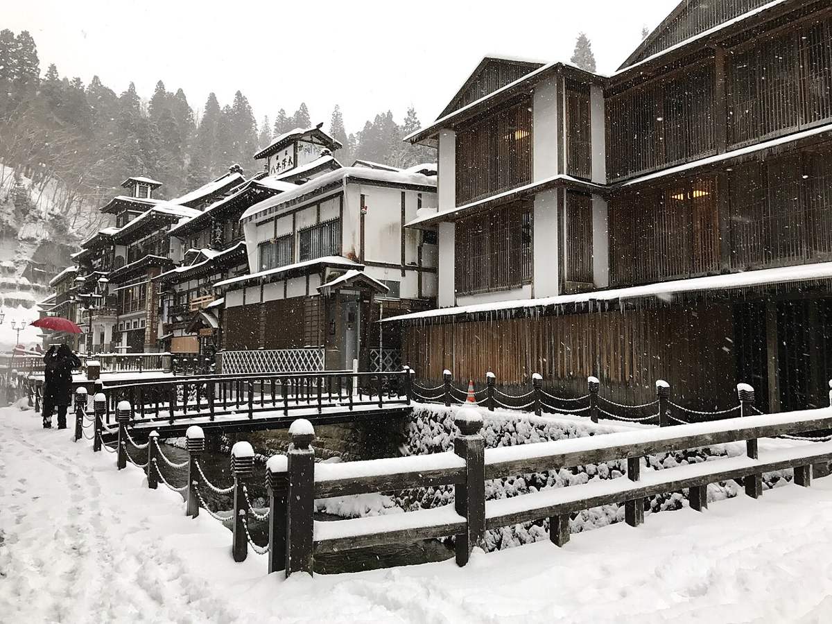 下雪中的銀山溫泉街步道:地面是被踩實的雪、留著一道道腳印,河岸鐵鍊欄杆與對岸旅館的木格柵全被雪蓋住,左後方一個撐紅傘的人背對鏡頭走著