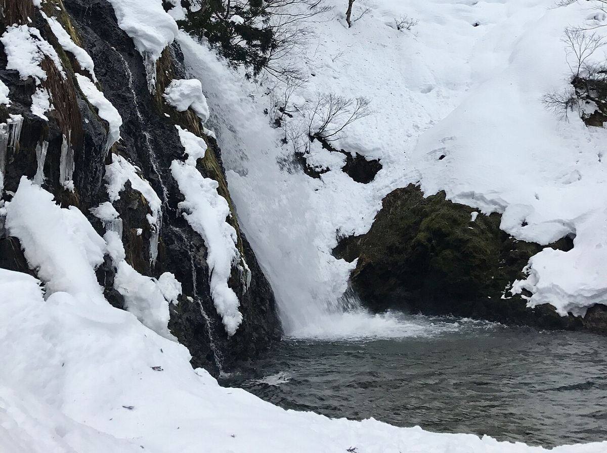 冬天的白銀之瀧:水流從左側黑色岩壁落進深色水潭,岩壁掛著冰柱、岩頂與右側斜坡覆蓋厚雪,前景的雪地滿是腳印