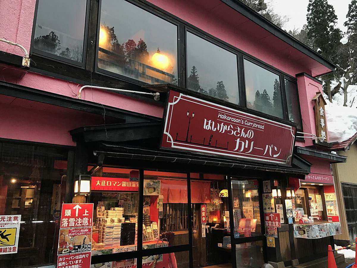 銀山溫泉街上一間粉紅色外牆的店面,門口立著紅底白字的「頭上 落雪注意!」告示牌,左邊另有黃底的「凍結注意」滑倒圖示牌,屋簷與後方山坡都積著雪