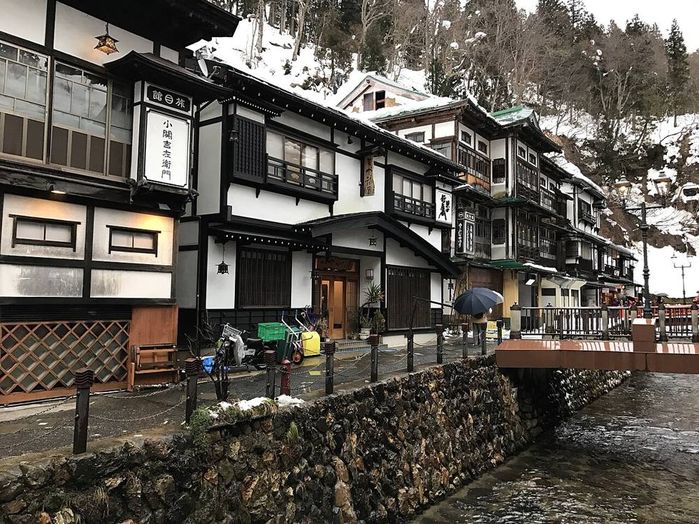 銀山溫泉街旁的木造旅館「小関吉左衛門」門口,停著一台送貨機車、兩台手推台車和幾個綠色塑膠箱,右前方是紅欄杆小橋與銀山川