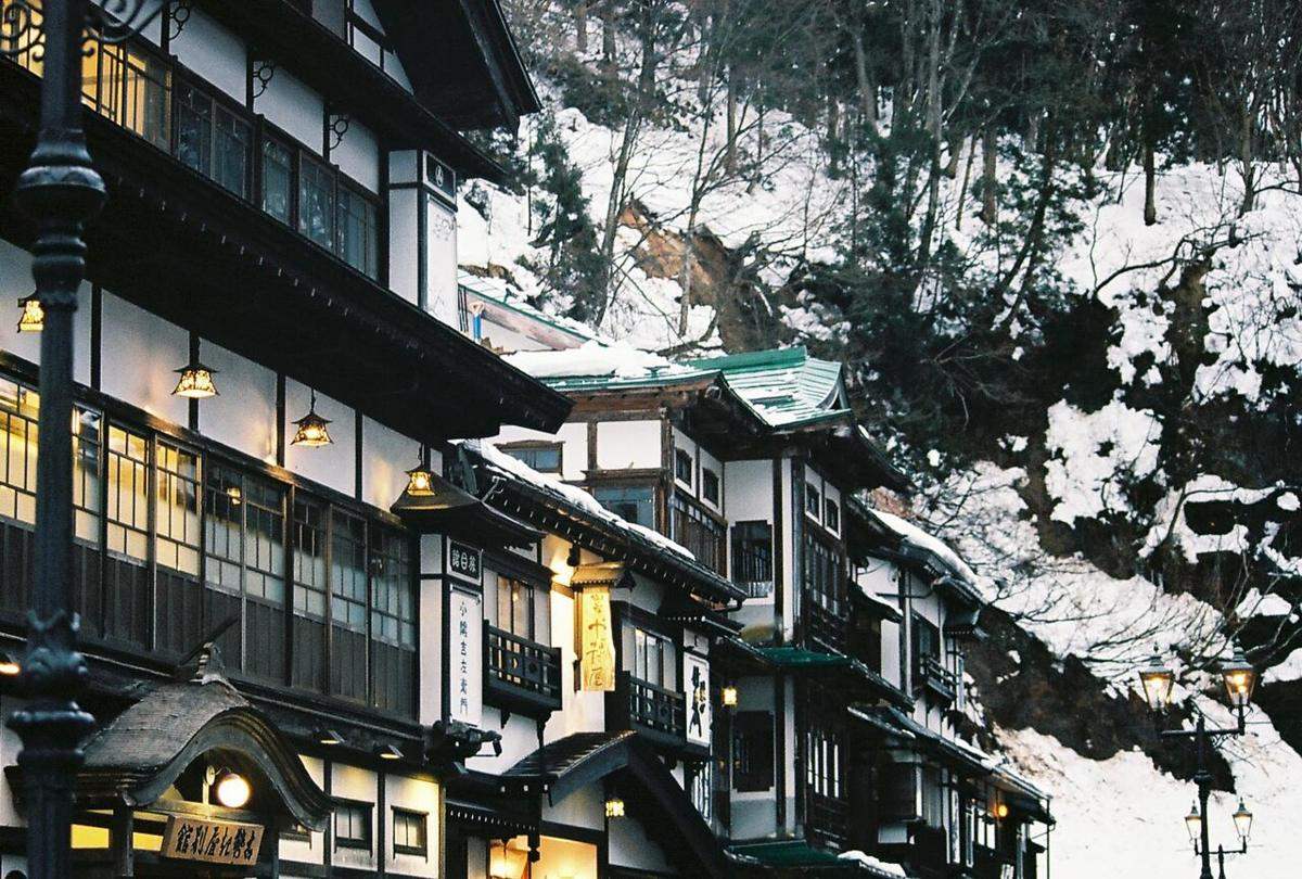 天色轉暗的銀山溫泉街:木造旅館二樓走廊的吊燈與房間窗戶亮著暖黃光,畫面左緣和右側各有一盞造型煤氣燈已點亮,背後山坡全是積雪