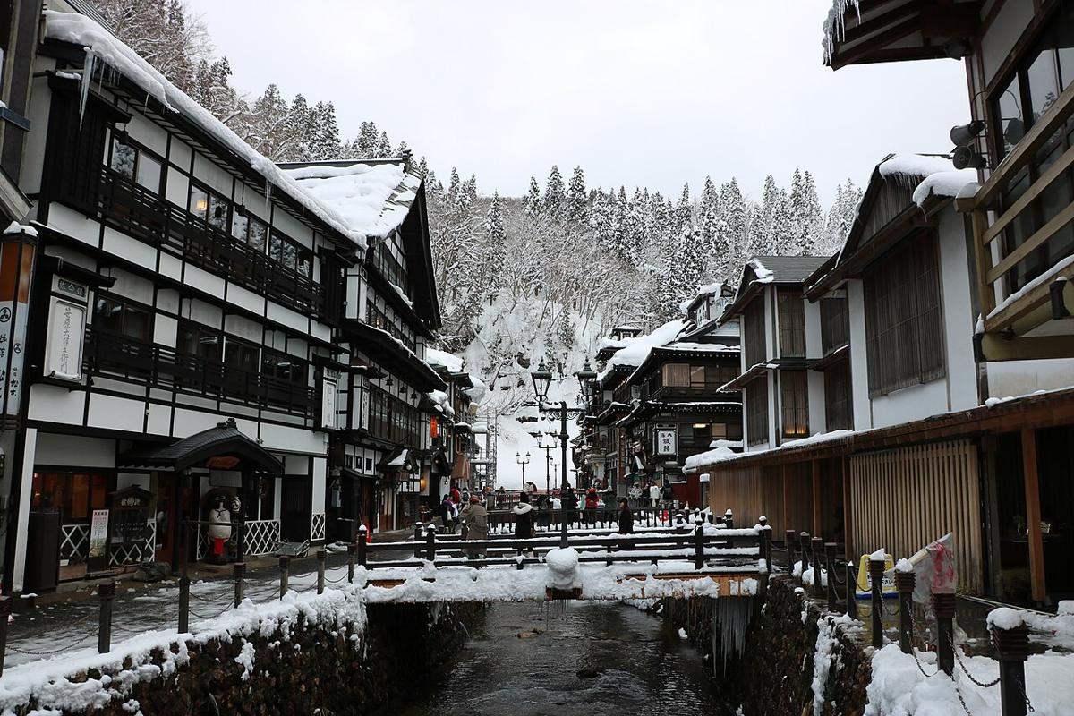 下雪後的銀山溫泉街:銀山川兩側木造旅館屋簷厚厚積雪,兩岸各只有一條窄窄的行人道,遠處小橋上有幾個遊客,路面上沒有任何車輛