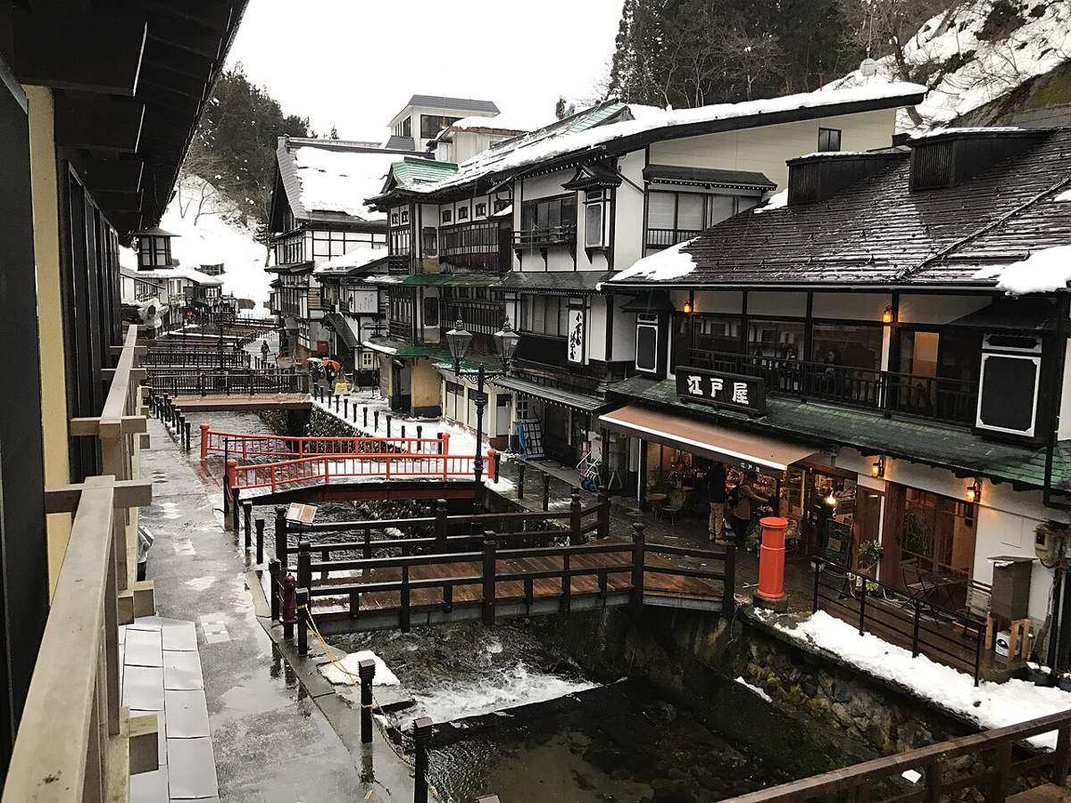 從旅館二樓外廊俯看銀山溫泉街:銀山川夾在兩排木造旅館中間,河上有一座紅欄杆小橋與兩座木橋,右側「江戸屋」招牌下是一排店面,屋頂與河岸殘雪未化