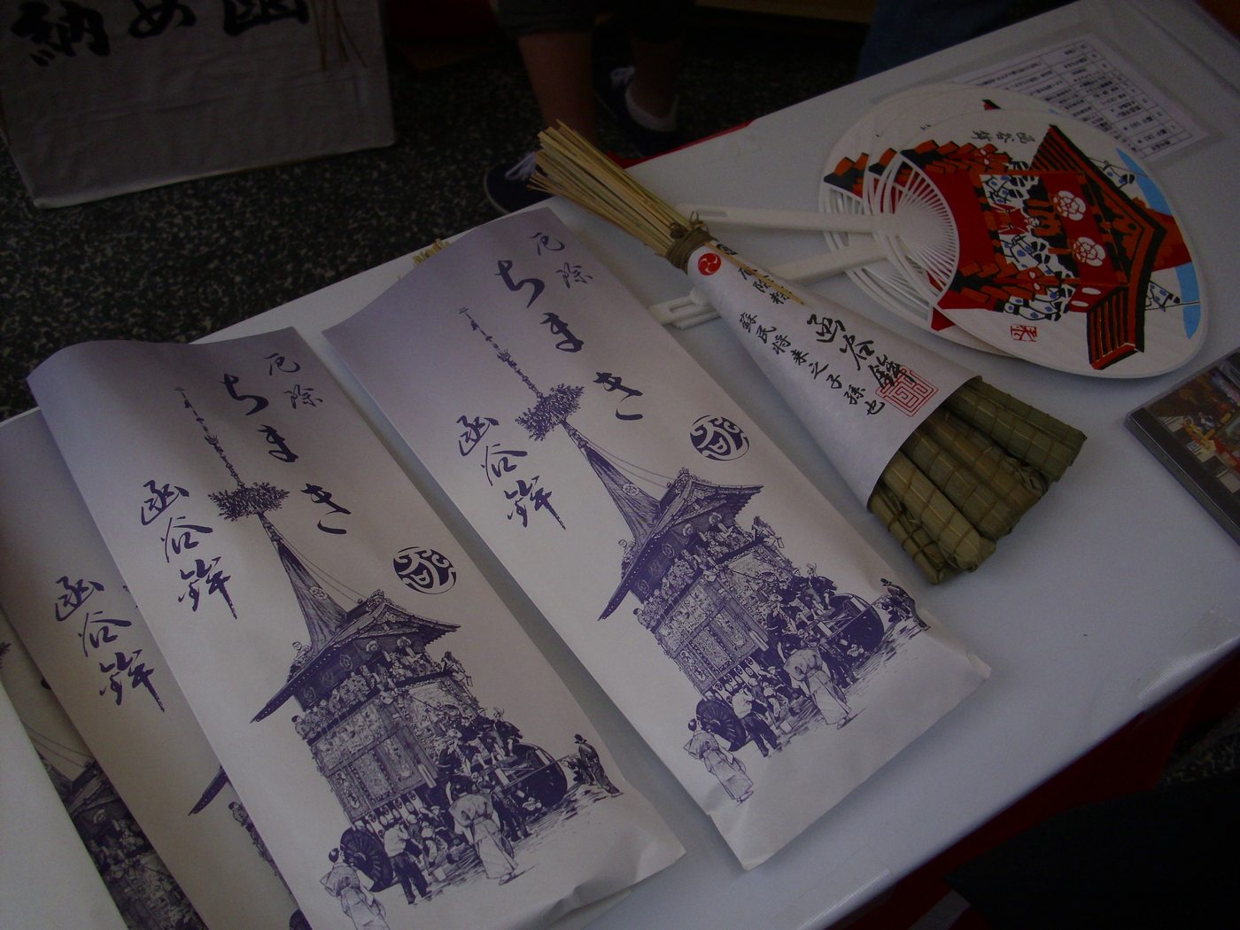 Long paper sleeves printed with the words for protective chimaki and the name Kanko-hoko laid out on a white cloth table, with a bundle of bound bamboo leaves showing out of one sleeve and paper fans beside them