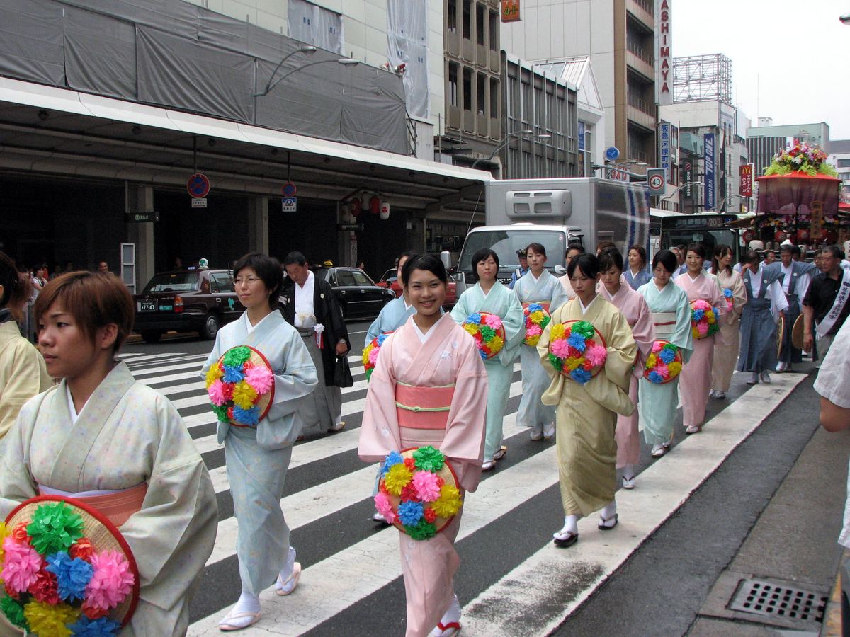 A line of women in kimono carrying flat straw hats covered in coloured paper flowers across the crossing at Shijo-Kawaramachi, with a flower-topped cart following behind on the right